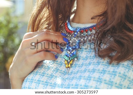 Brown-haired girl in a blue dress with a necklace. Large necklace rhinestones. Thin hand with blue nail. Fashion trends. Street fashion. Trendy, stylish. Fashionable girl.