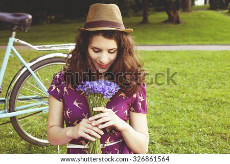 Beautiful brunette with a bouquet of cornflowers. Girl with retro bicycle. Girl in a park on the lawn. Beautiful girl with curly hair. Young girl in  purple dress and hat. Inhale the scent of flowers.
