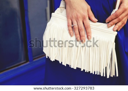 Hands and white bag with fringe. White bag with fringe. Accessories. Fashion handbags. Tanned hands. A young woman, girl. Hands-on bag. The blue skirt and white bag.