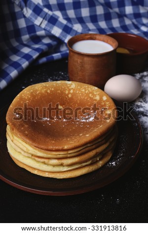 Stack of pancakes, flour, eggs and milk on the table. Cooking pancakes. Napkins, tablecloths. Kitchen table, cooking. Pancakes on a clay plate. Low key. Clay cup with milk, eggs and pancakes.