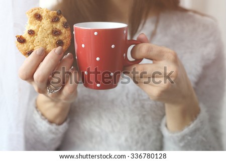The girl in a sweater with a cup of tea or coffee in their hands. Coffee and biscuits. Cup and cookies with raisins. Bitten cookie. Hold the cup in his hands. Warming hands.  Drink. Breakfast.