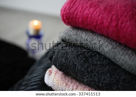 A stack of woolen sweater on the bed. Gray, pink and black sweater. Warm and cozy. Knitted clothes. Burning candle and warm clothing. Home atmosphere, winter clothing.