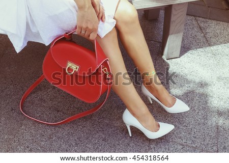 Young woman in white summer classic leather high heel shoes with bright red leather handbag. Girl in a white dress with glitter tattoo on her leg resting on the summer terrace. Handbag fashion look