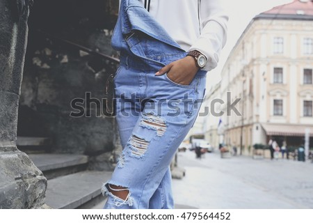 Woman in ragged blue denim overall posing on the city streets. Stylish girl in trendy overall, spring casual fashion look.