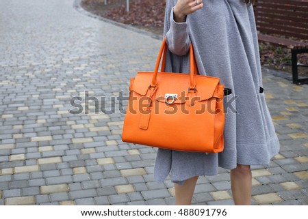 Autumn street fashion look. Young brunette girl in a gray knitted long dress with large bright orange leather bag in hand in the park.