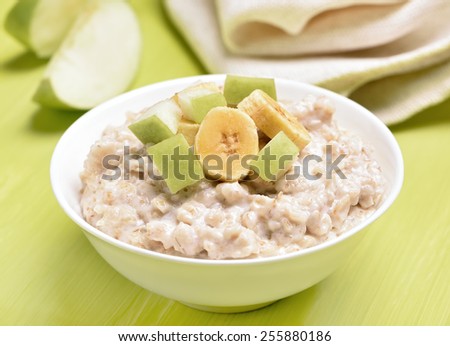 Oatmeal porridge with apple and bananas slices in white bowl on green table