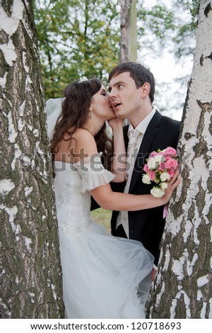 Handsome bride and funny groom stands in the park between trees. Bride whisper something to groom.