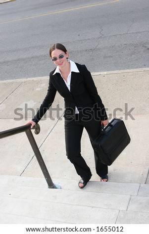 Young woman in business attire, carrying briefcase and smiling with one foot on some steps.