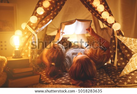 happy family mother and children reading a book with a flashlight in a tent at home