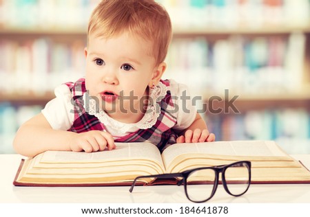 Happy funny baby girl in glasses reading a book in a library