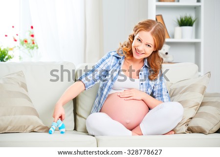 happy pregnant woman is resting at home on the sofa and holding a little blue booties socks