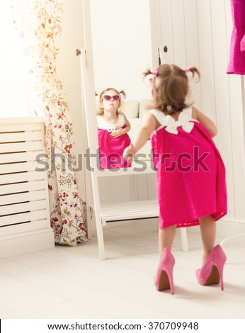little girl child fashionista looking in the mirror at home in a pink dress, shoes and sunglasses