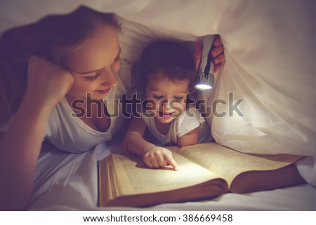 Family reading bedtime. Mom and child daughter reading a book with a flashlight under the blanket in bed