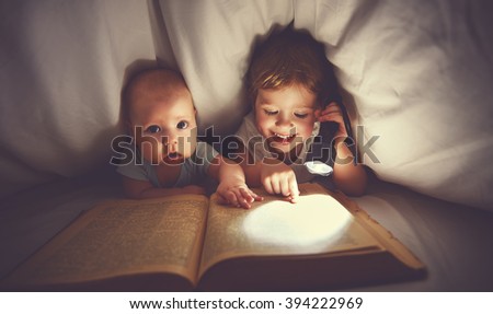 children brother and sister read a book with flashlight under blanket in bed