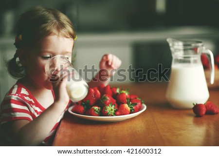 happy child girl drinks milk and eats strawberries in the summer home kitchen