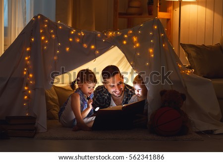 happy family father and children reading a book with a flashlight in a tent at home