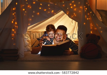 happy family father and child daughter  reading a book with a flashlight in a tent at home