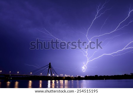 Flash in dark sky near the bridge in the night. Lightning like a tree. lightning clouds, and reflection of lights on the river\'s water