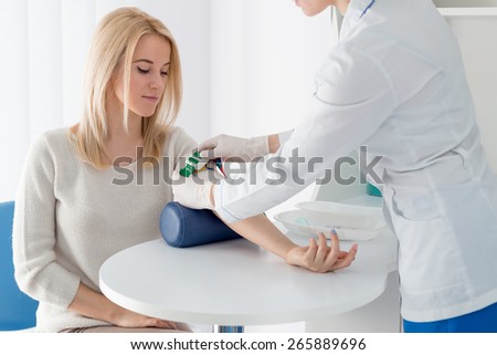 Preparation for blood test with beautiful young blond woman by female doctor in white coat medical uniform on the table in white bright room. Nurse patient hand grips green jute.