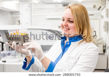 Nurse woman in white uniform and gloves looking at the stand with the test tubes with blood in the light medical laboratory.