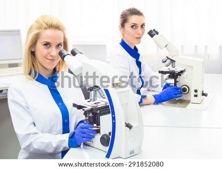 Two medical workers women in white uniforms and gloves working with microscope that is on the table in light and white medical lab. Nurse pretty blonde and brunette.