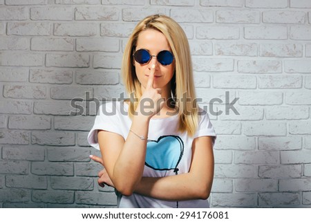 Beautiful blonde girl in branded sunglasses smiling in studio front of a white brick wall. Dressed in a white t-shirt.