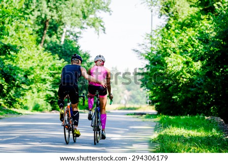 Beautiful young pair of professional cyclists riding on the road cose to each other hugs surrounded by green trees on a bright and sunny day.Girl in pink jumpsuit with a helmet,guy in a dark blue suit