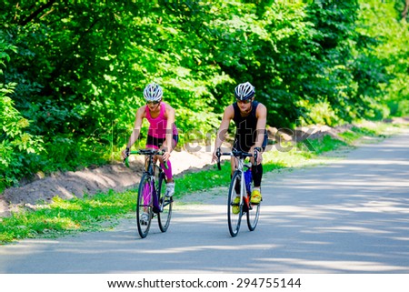 Beautiful young pair of professional cyclists in gear and glasses riding on the road surrounded by green trees on bright sunny day. Girl in pink jumpsuit with helmet, guy in dark blue special suit.