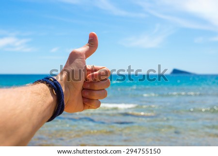 Close-up picture of mans hand with rope bracelet on the beach against a blue background and the blue sea with waves and a mountain. Hand showing thumbs up or shows good.
