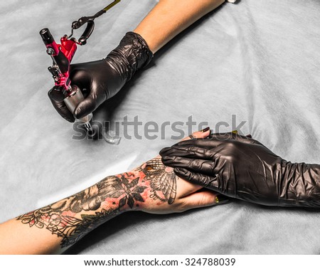 Tattoo master take paint in salon for tattoo on girls hand. Tattooist works in professional salon with pink tattoo machine on a blue mat and in sterile black gloves.