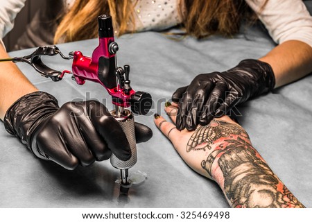Tattoo artist takes an orange paint for black and red tattoo which draws for a girl. Master works in a professional salon with pink tattoo machine on a blue mat and in sterile  black gloves.