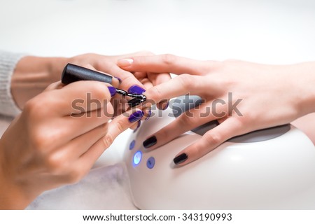 Closeup manicurist applies nail gel polish on middle finger. Girl client holds hand on the client on Uv lamp. Beautician gently manicured service.