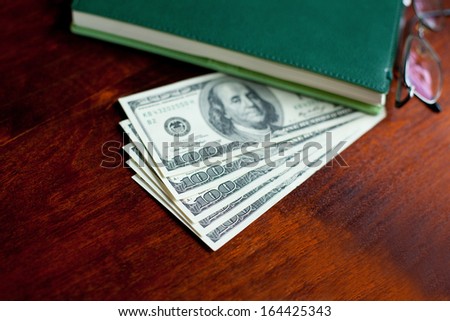 Stacks of one hundred dollars banknotes, notebook, pen and glasses on wooden desk