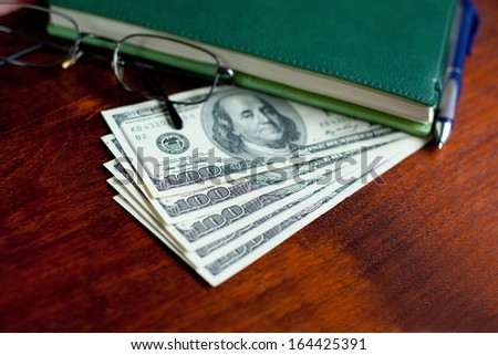 Stacks of one hundred dollars banknotes, notebook, pen and glasses on wooden desk