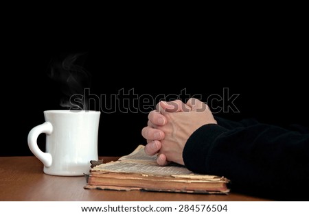 woman\'s hands folded in prayer on a worn vintage Bible with white coffee mug