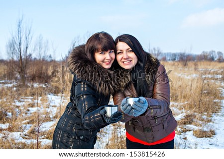 Two beautiful woman friends dressed in winter clothes enjoying a conversation, drinking hot drink and laughing together outdoors