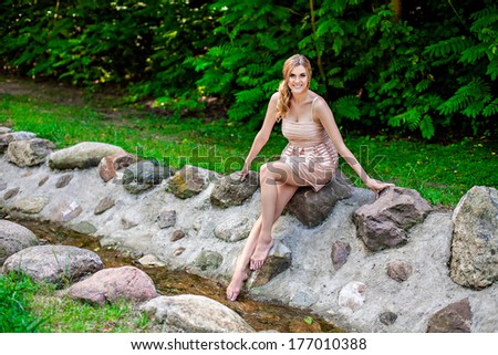 Young beautiful smiling woman sitting on rock by stream in park