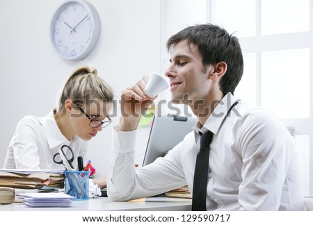 Business man enjoys a hot cup of coffee in the office, annoyed female colleague in the background