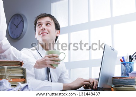 Hand of colleague offering coffee to young man sitting on workplace behind computer