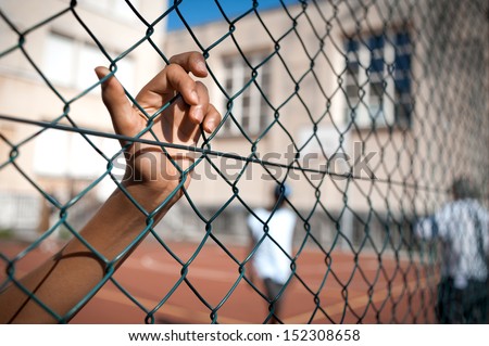 A boy\'s hand clinging on to a fence, basketball court on background