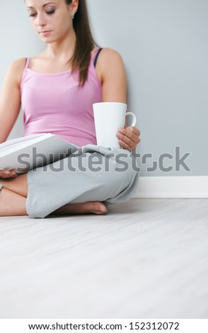 Young woman with book and a cup of coffee sitting on floor, focus on hand