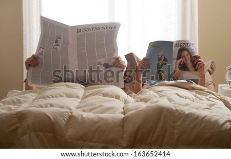 Young Couple In Bedroom Reading Newspaper And Magazine