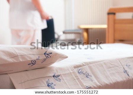 Bed at nursing home with nurse and wheelchair on background.