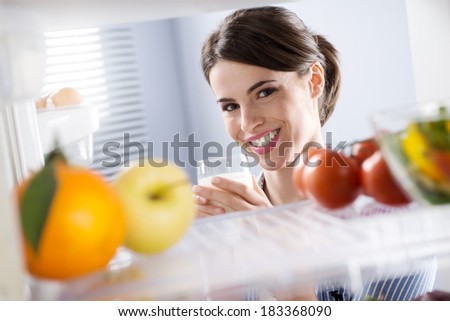 Attractive woman smiling and holding a glass of milk in front of refrigerator.