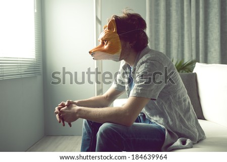 Young man wearing a fox mask sitting on sofa in front of a window.