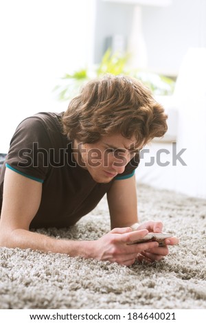 Young man with smartphone lying down on living room carpet.