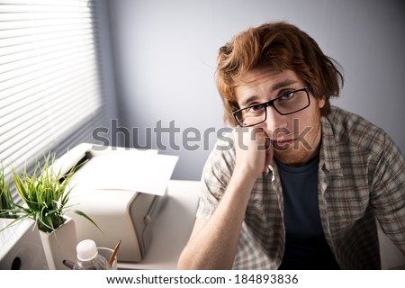 Young bored guy at office staring at camera with printer and plants on background.