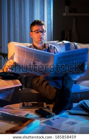 Businessman reading financial newspaper late at night sitting on sofa.