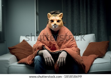 Young man wearing a fox mask sitting on sofa covered with a blanket.