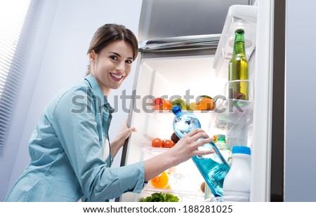 Young woman taking a water bottle from refrigerator and smiling at camera.